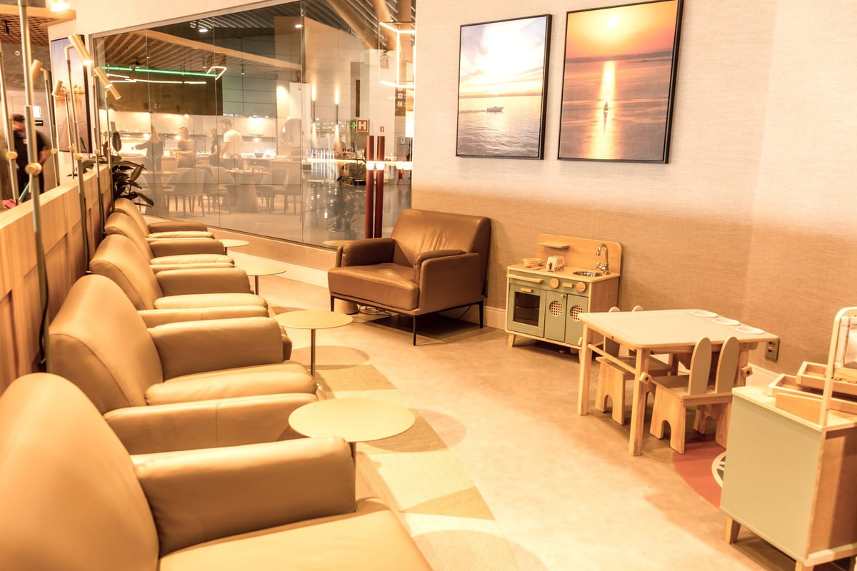 Seating area and children's play corner at W Premium Lounge in Porto Alegre Airport, featuring leather lounge chairs, side tables, framed sunset artwork, and a mini wooden play kitchen and furniture set for young travelers.