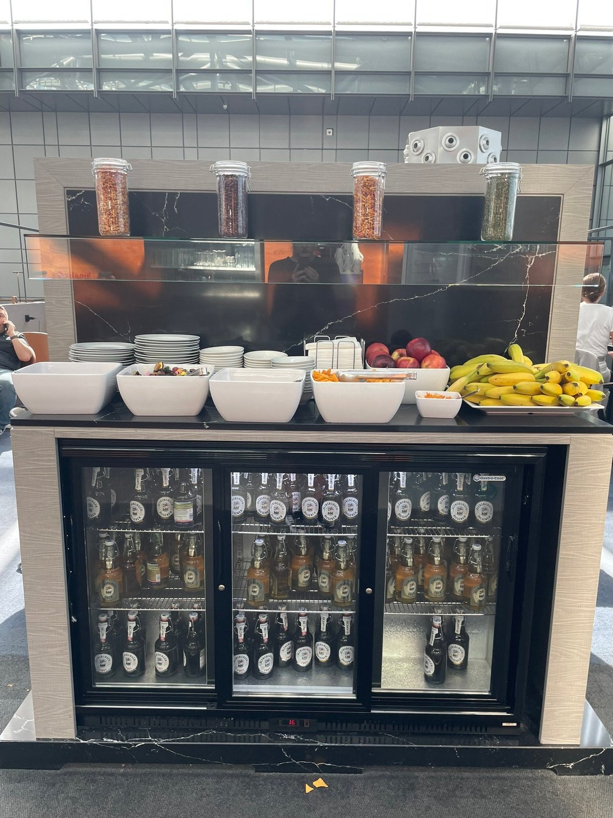 A well organised snack and beverage station at the Hamburg Airport Lounge. There is a refrigerator stocked with beer, while the countertop above displays several white bowls filled with fresh fruits, nuts and other snacks.