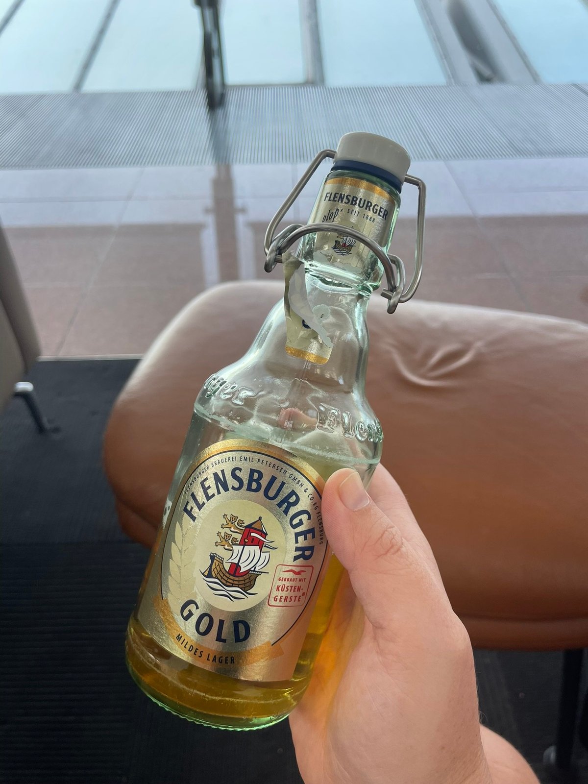 The image shows a person holding a bottle of Flensburger Gold, a mild lager beer. The bottle has a distinctive flip-top cap and a label featuring the brand's logo, which includes a ship and traditional German design elements.