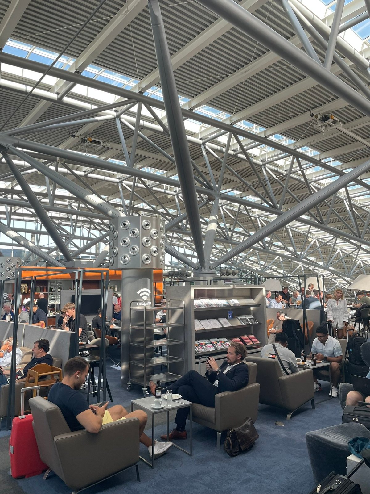 This image shows a bustling section of the Hamburg Airport Lounge, featuring passengers seated in comfortable armchairs, some enjoying snacks or drinks while others use their phones or work. In the centre is a magazine and newspaper stand.