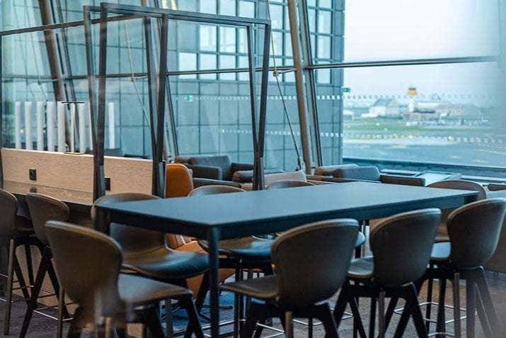 A modern seating area at Hamburg Airport Lounge, featuring high tables and bar style chairs. The dark furniture creates a sleek and functional workplace or dining area for travellers. Large floor to ceiling windows in the background offer views.