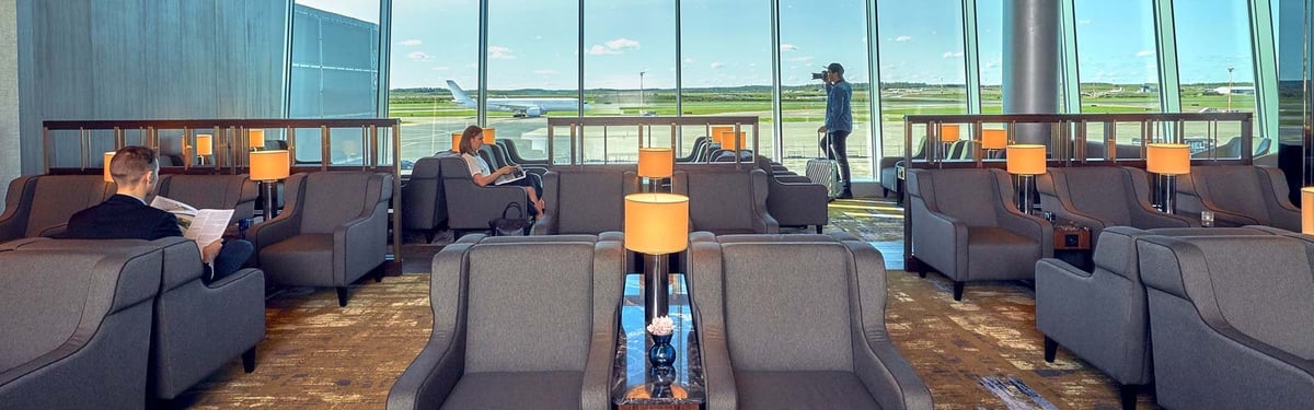 Interior view of the Plaza Premium Lounge at Helsinki Airport, featuring a spacious seating area with comfortable grey armchairs and side tables topped with modern lights. Large windows provide natural light and a clear view of the tarmac.