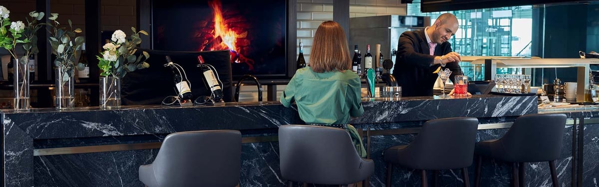 Bar area of the Plaza Premium Lounge at Helsinki Airport, featuring a sleek black marble countertop adorned with elegant floral arrangements in vases. A bartender in a suit prepares a cocktail while a guest sits at the counter.