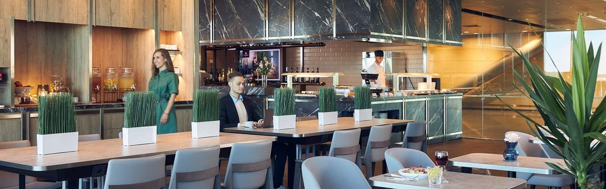 Dining area of the Plaza Premium Lounge at Helsinki Airport, featuring a modern design with communal and individual tables. Tables are decorated with minimalist greenery in sleek white planters. There is a buffet station in the background.
