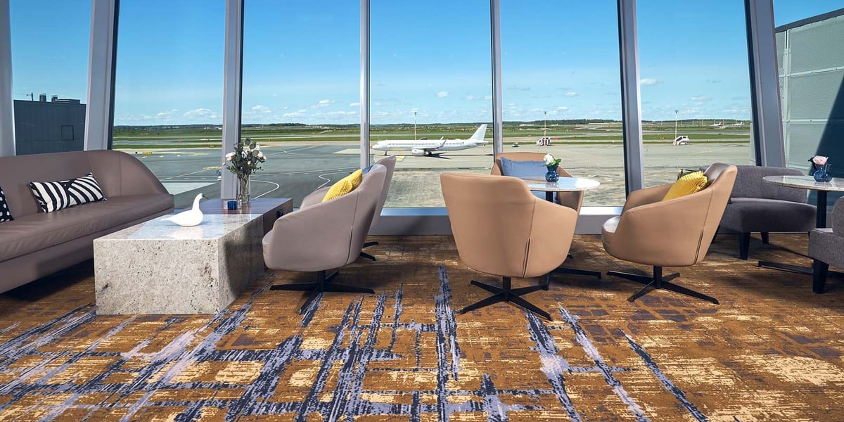 View of the Plaza Premium Lounge at Helsinki Airport, showcasing a comfortable seating area with stylish chairs, sofas, and marble-top tables. The large floor-to-ceiling windows provide a clear view of the airport tarmac where planes are visible.