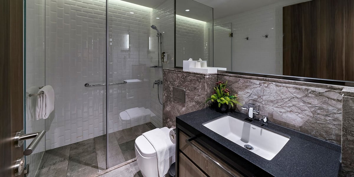 Shower room at the gate1 Plaza Premium Lounge at Hong Kong Airport. The shower has white subway tiles on the walls and a rainfall showerhead. The vanity area has a dark countertop, white sink, a large mirror and a decorative arrangement of greenery.