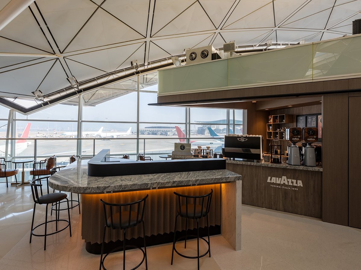 This image shows a modern and stylish coffee bar at the gate 60 Hong Kong plaza premium lounge, with large window views of the tarmac and planes in the background. The bar has a sleek marble countertop with a few barstools for seating.