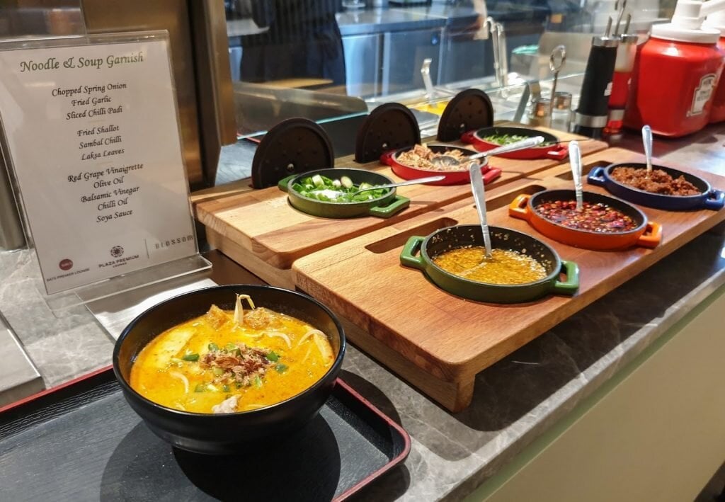 Noodle and soup station at Blossom Lounge Changi Airport Terminal 4 with laksa bowl and condiments including sambal, fried garlic, and chili.