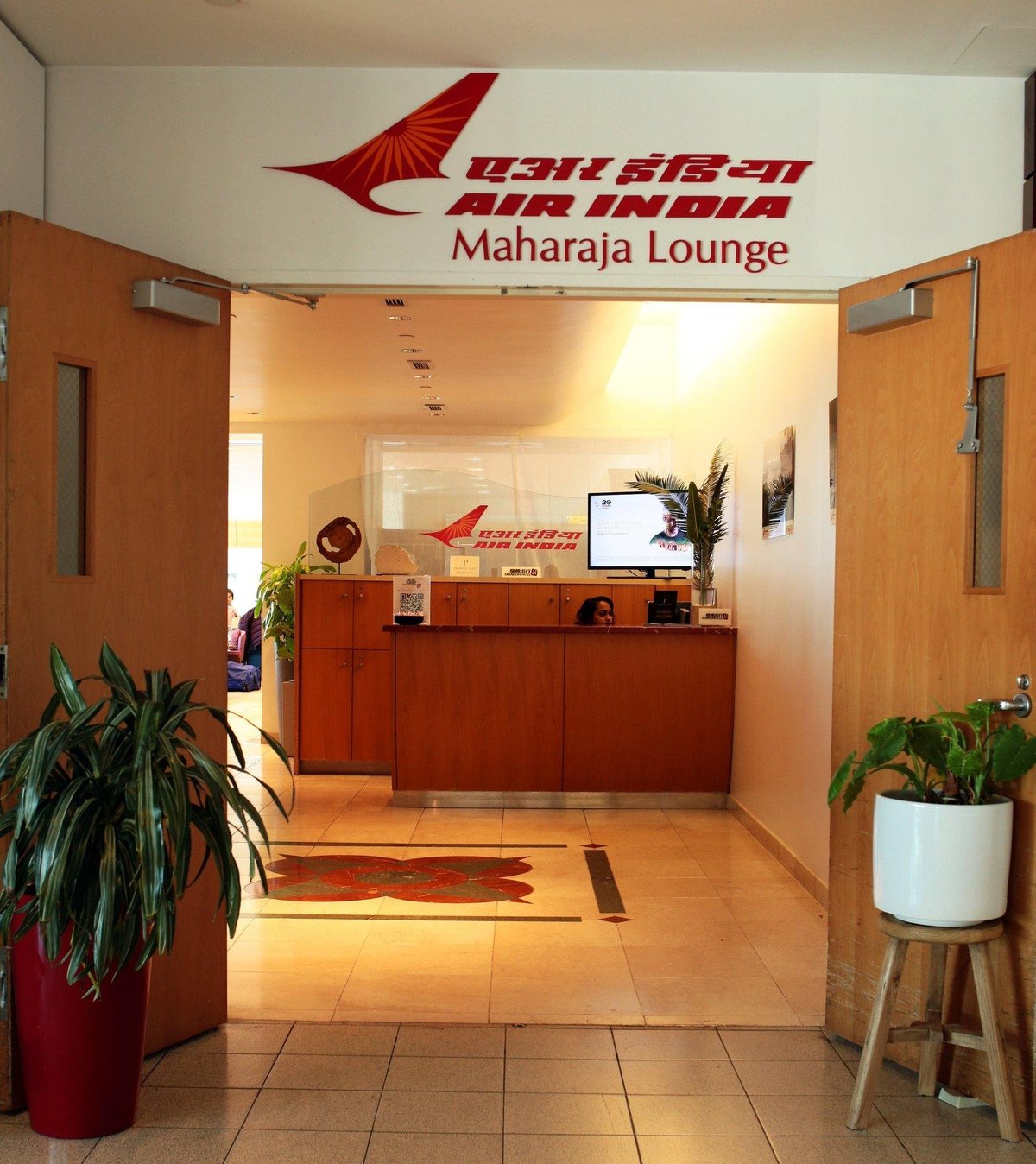 Entrance of the Air India Maharaja Lounge with the airline's logo and name prominently displayed above the reception desk. The floor is adorned with a branded carpet, and the area is accented with green potted plants, creating a welcoming atmosphere