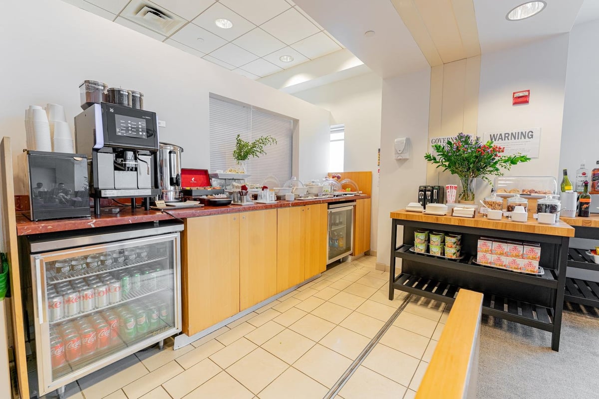 The buffet section of the Air India Maharaja Lounge featuring an espresso machine, soft drinks and a selection of snacks displayed on the counter. A well organised area that includes condiments and neatly arranged shelves with packaged goods.