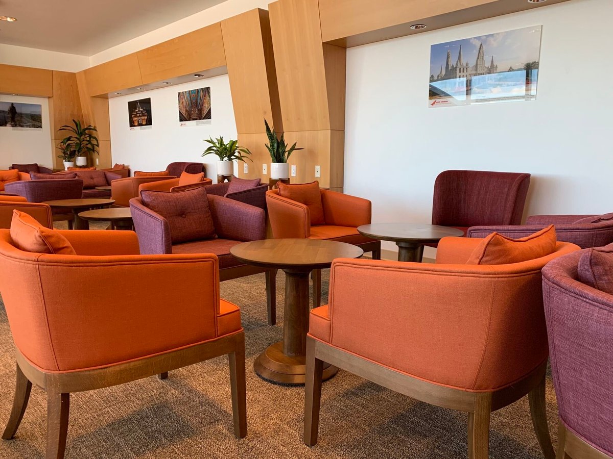 The Air India Maharaja Lounge at JFK features a mix of orange and maroon armchairs clustered around wooden tables. The room is bright with natural light and adorned with wall art and potted plants, offering a comfortable space for passengers to relax