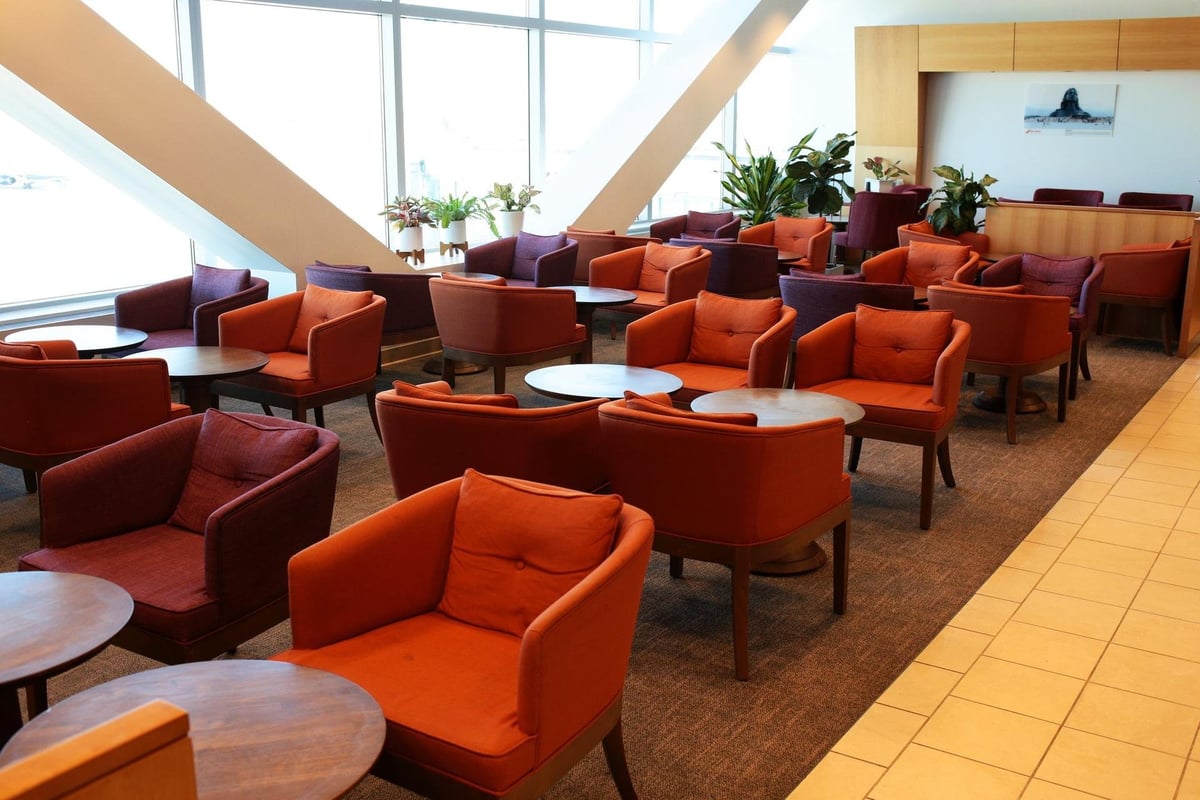 Air India Lounge JFK with red and orange armchairs arranged around circular tables, under natural light streaming from large windows. The lounge features a warm, welcoming atmosphere, with plants and a picture on the wall.