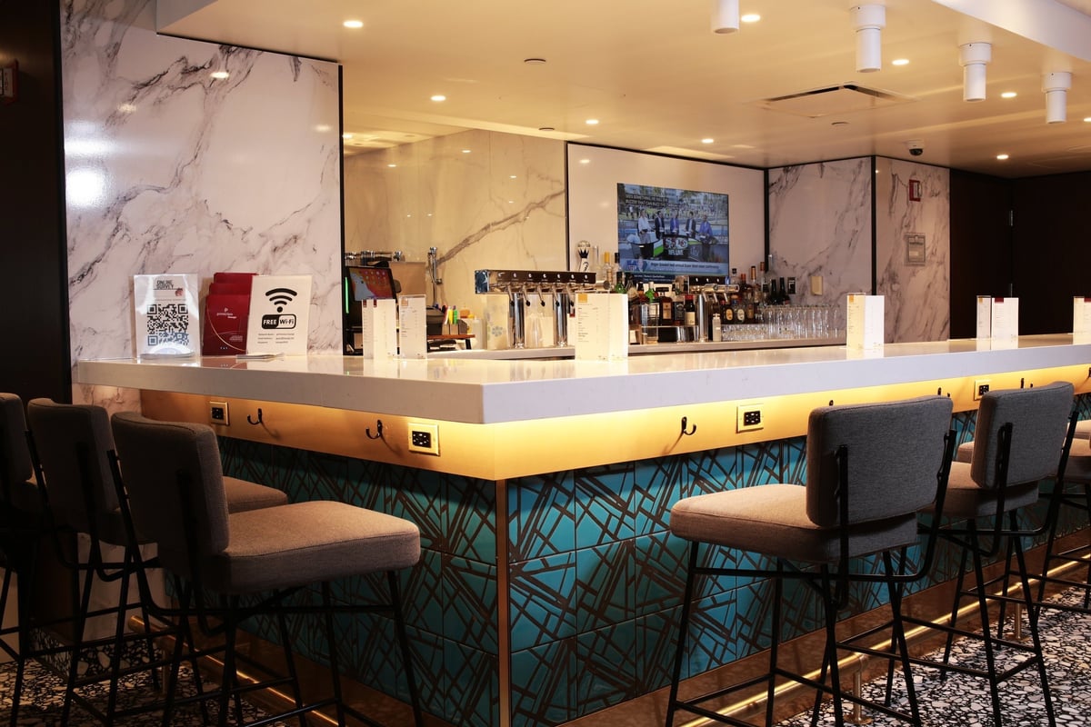 Bar seating area at JFK Primeclass Lounge T1 featuring a well-stocked bar with a marbled or marbled appearance back wall with an embedded tv. The bar has several barstools and menus along its countertop, as well as other signs such as for wifi.