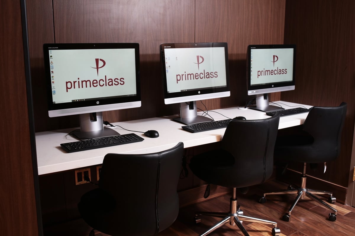 Business computers positioned on a white desk lined with black office chairs at the JFK T1 Priceless Lounge.