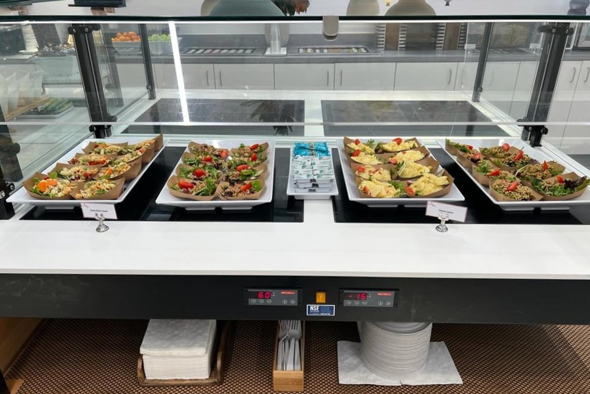 A food display at the Primeclass Lounge in JFK's Terminal 4, offering a selection of salads, sandwiches, and refreshments. The clean, modern buffet setup is well-maintained, with trays of food neatly presented under a glass cover and utensils.