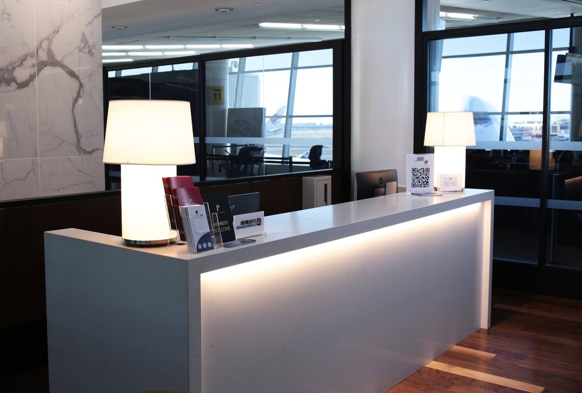 Primeclass Lounge T1 JFK airport front desk reception area with wide window views of the tarmac and bright lamps on either side of side of the desk. Informational pamphlets are positioned on the desk.