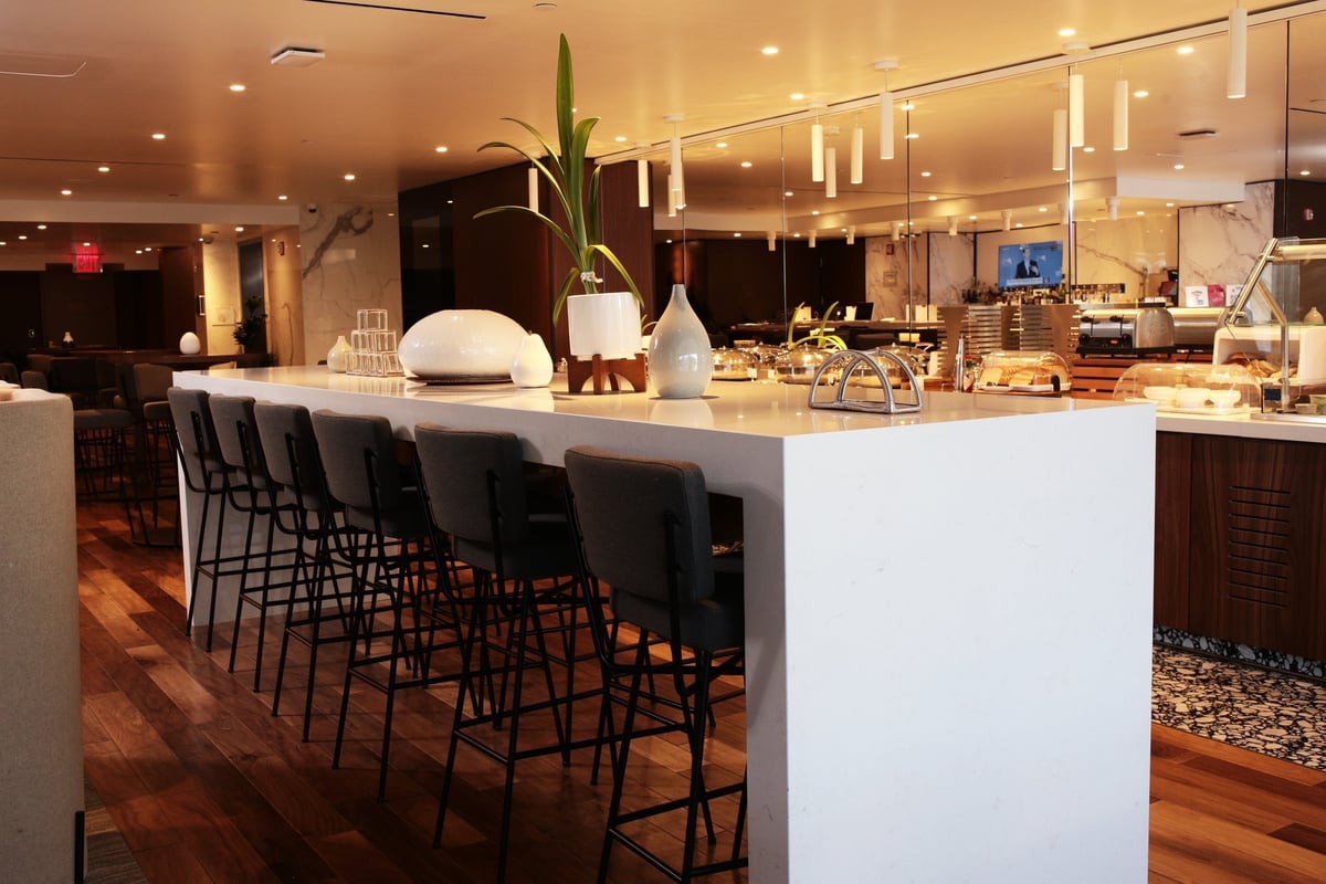 Hightop table seating Primeclass Lounge T1 JFK featuring seating area next to a buffet with high stools and a decorative plant in the centre. The buffet station includes various items under food plates and a beverage station.