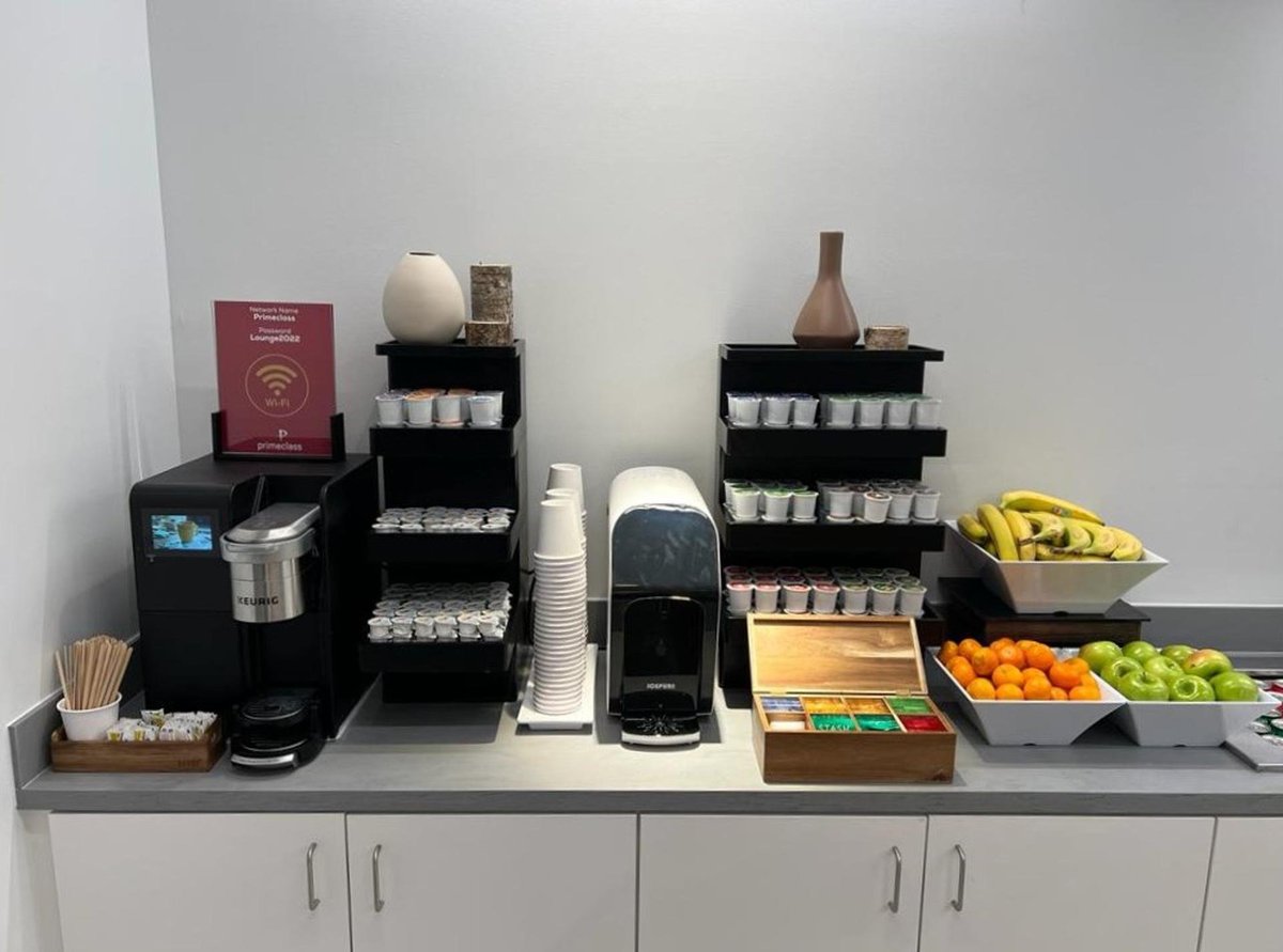 A well-organized coffee and tea station inside the Primeclass Lounge at JFK's Terminal 4, featuring a Keurig machine, an assortment of tea bags, and fresh fruit. The station is neatly arranged with an array of cups and condiments.