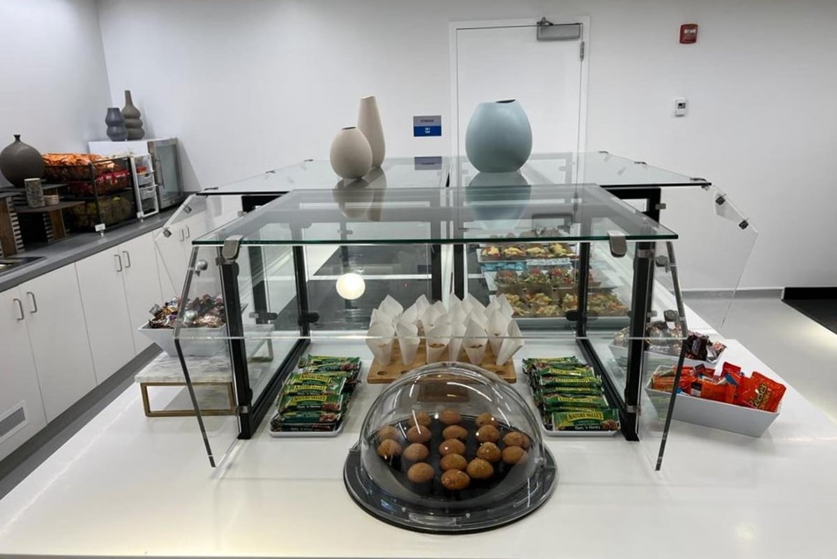 Buffet counter inside the Primeclass Lounge at Terminal 4 of JFK Airport, showcasing an array of snacks including muffins and packaged nuts under a glass display. The countertop is clean and white, contributing to a sleep design.