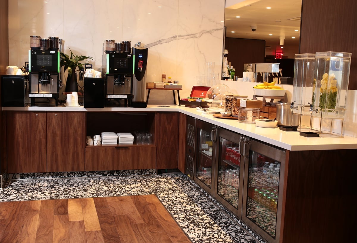 Beverage station at the T1 Primeclass Lounge at JFK Airport, featuring a coffee machine, various condiments, a small fridge at the bottom with various drinks including soft drinks, and a fruit basket on the countertop.