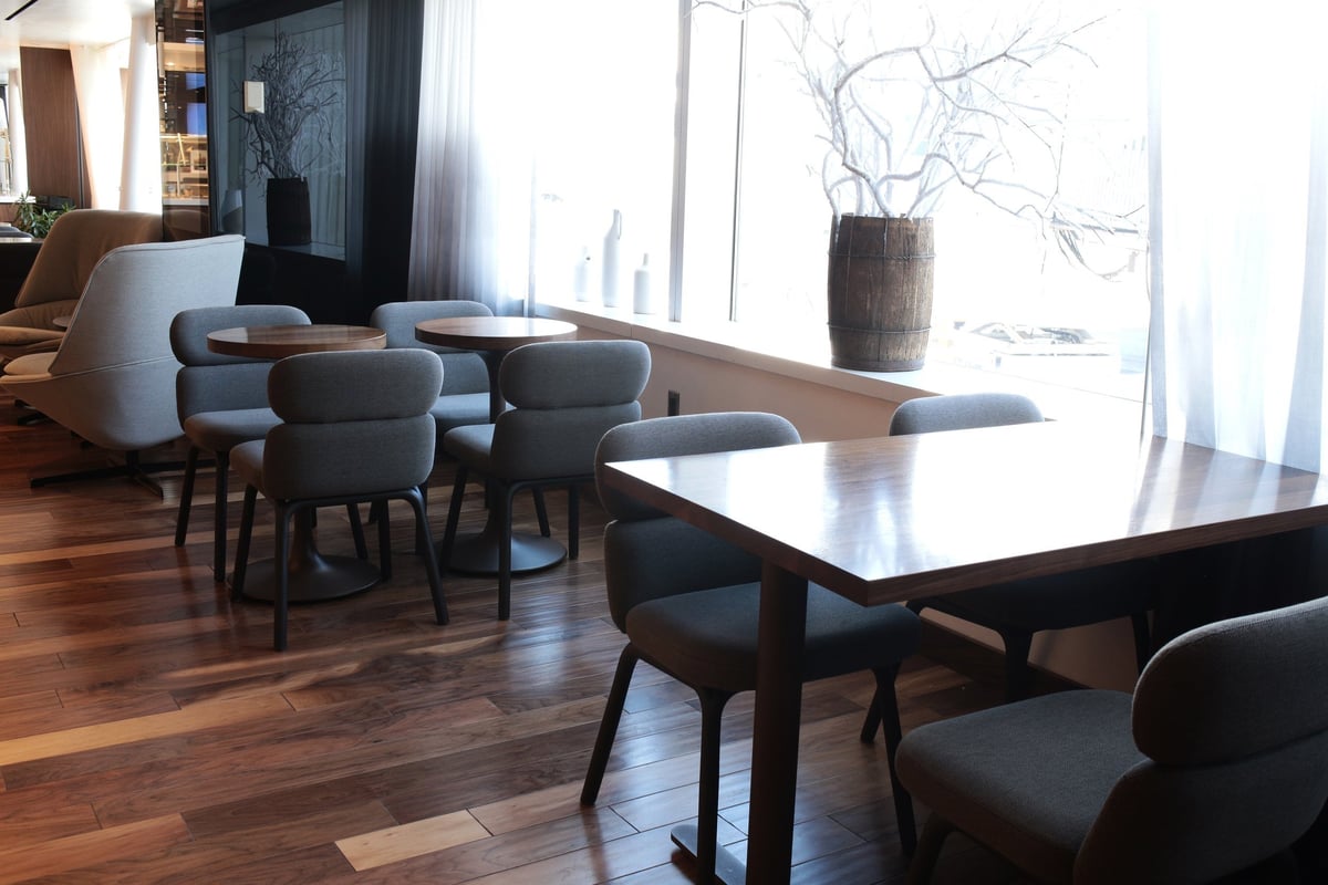 Seating area near a window at the T1 Primeclass Lounge JFK Airport. Tables and chairs of 4 are seated against the side of the room next to a bright window with a decorative planet in a wooden barrel on the ledge.