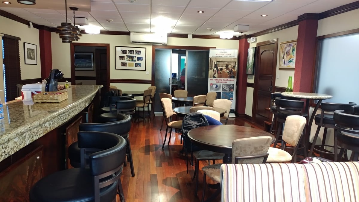 Club Kingston Arrivals Lounge at Norman Manley International Airport featuring a granite-topped bar, round dining tables with cushioned chairs, wood flooring, and promotional signage for cardholder access