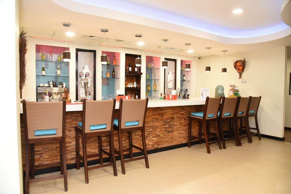Modern airport lounge bar at Club Kingston in Norman Manley International Airport with wicker high chairs, illuminated shelves of liquor bottles, and decorative Caribbean wall art