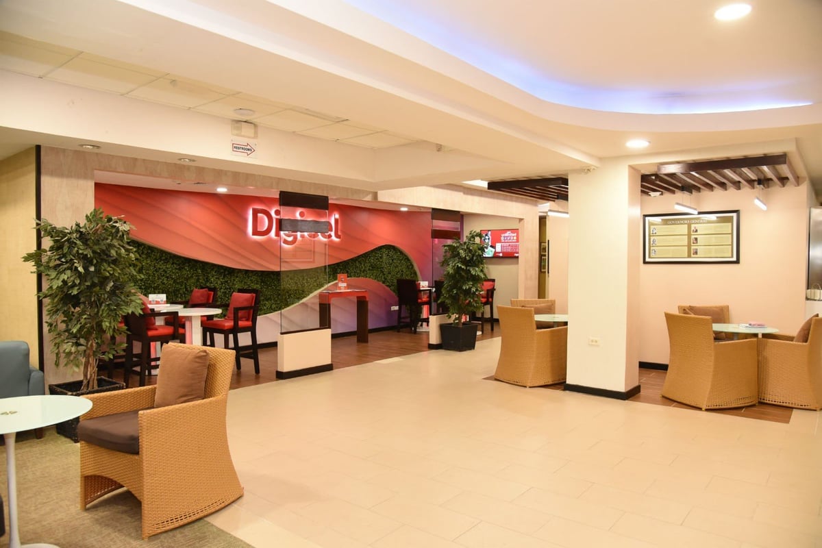 Open seating area in Club Kingston departure lounge at Norman Manley International Airport featuring Digicel-branded workspace, wicker chairs with cushions, modern lighting, and indoor greenery