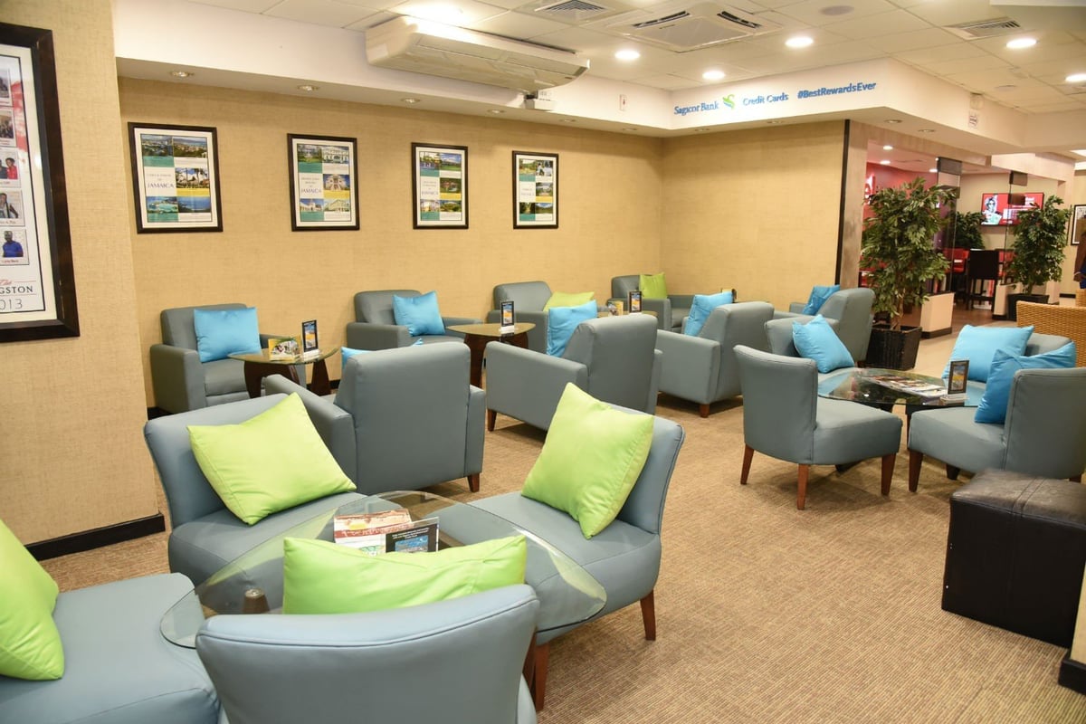Comfortable seating area in Club Kingston departure lounge at Norman Manley International Airport featuring cushioned armchairs with bright green and blue pillows, wall art, and ambient lighting