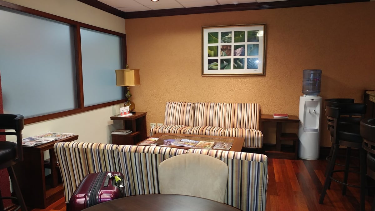 Cozy seating corner in Club Kingston Arrivals Lounge at Norman Manley International Airport, featuring striped sofas, a wooden coffee table with magazines, a water dispenser, and soft ambient lighting