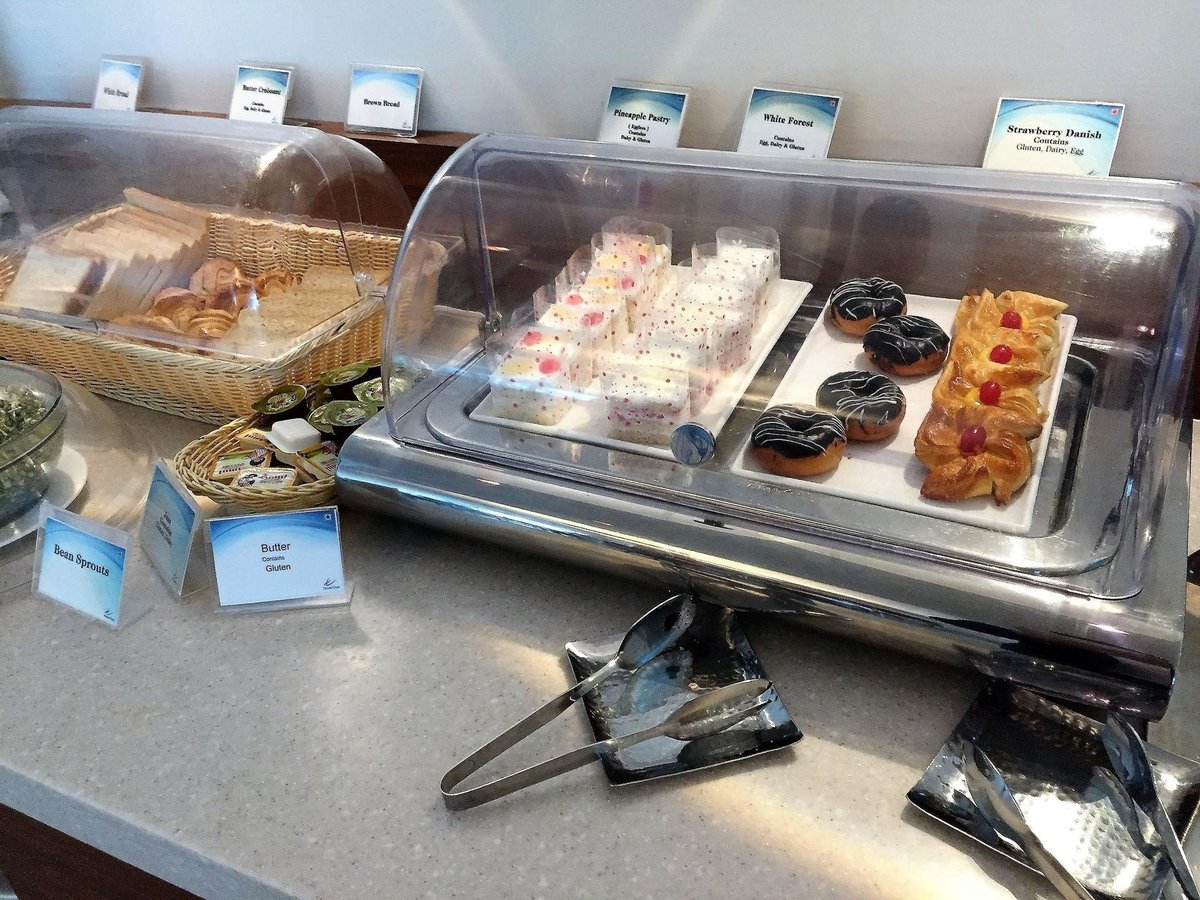 Buffet at T2 Travel Club Lounge Kolkata Airport. Close up of food under transparent lids are featured, including strawberry danish pastries, bread, white forest deserts, butter and bean sprouts. Tongs are positioned beside the containers.