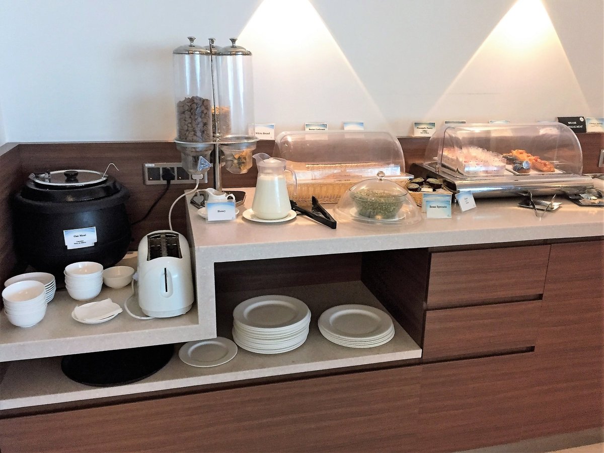 Buffet station at the Travel Club Lounge T2 Intl Kolkata Airport. The image showcases a buffet section with various food items such as pastries under transparent lids. There is a pitcher of milk, a toaster and several stacks of plates and bowls.