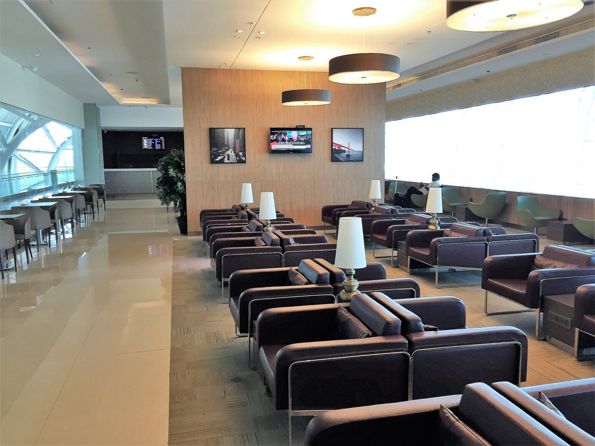 Seating area at the Travel Club Lounge t2 International Kolkata Airport. The image shows two rows of back to back leather burgundy seating with lamps in between them. To the left and right of the room lined up against the windows are green chairs.