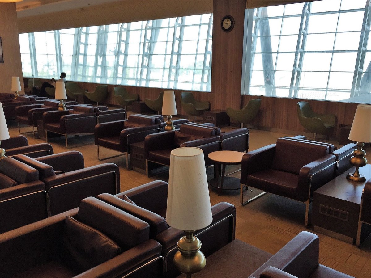 Travel club lounge at t2 international Kolkata Airport, featuring a seating area in the lounge with leather upholstered chairs in brown in the foreground and in green in the background against generous windows.