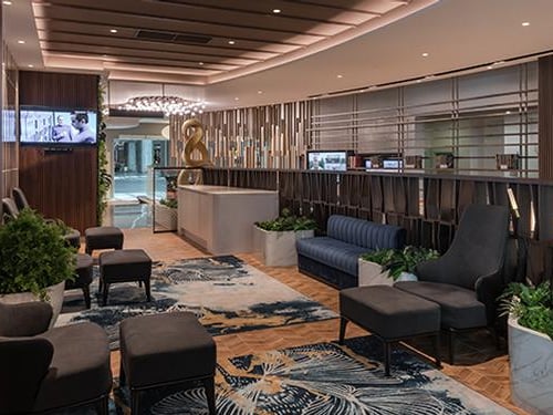 Reception and waiting area in the Travel Club Lounge at KLIA Terminal 2, featuring elegant seating, patterned rugs, lush greenery, and soft lighting with a modern chandelier and sleek wooden ceiling panels.