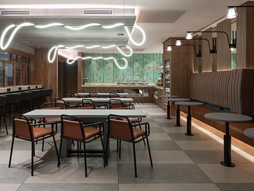 Modern dining area inside the Travel Club Lounge at KLIA Terminal 2, featuring tiled floors, minimalist tables and chairs, neon wave lighting fixtures, and a pastel green buffet wall with floral accents.