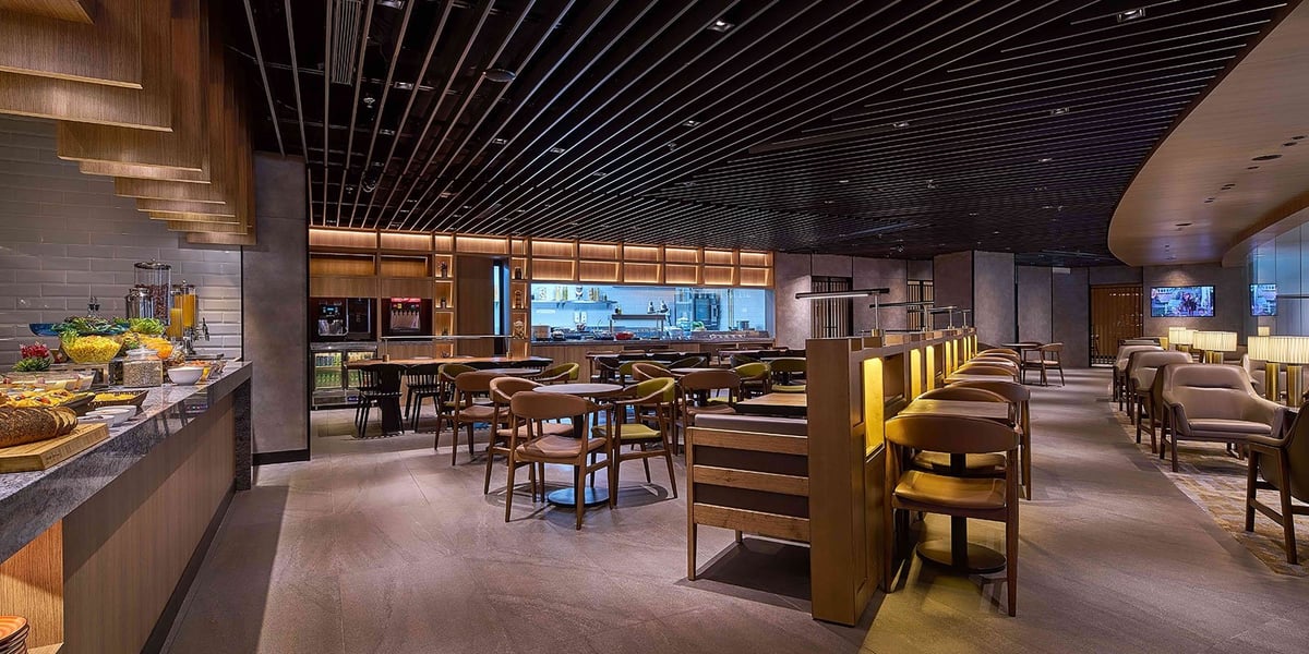Modern dining area inside the Plaza Premium Lounge at KLIA Terminal 2, featuring a well-lit buffet with fresh food options, a sleek open kitchen, and a mix of café-style tables and leather lounge chairs under a dark slatted ceiling.