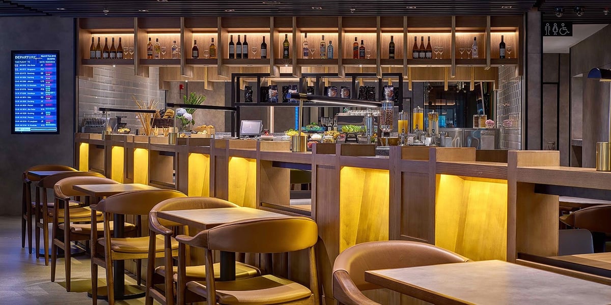 Bar and buffet area in the Plaza Premium Lounge at KLIA Terminal 2, featuring high-top seating, illuminated wooden paneling, a selection of alcoholic beverages, fresh juices, and a digital departure board.