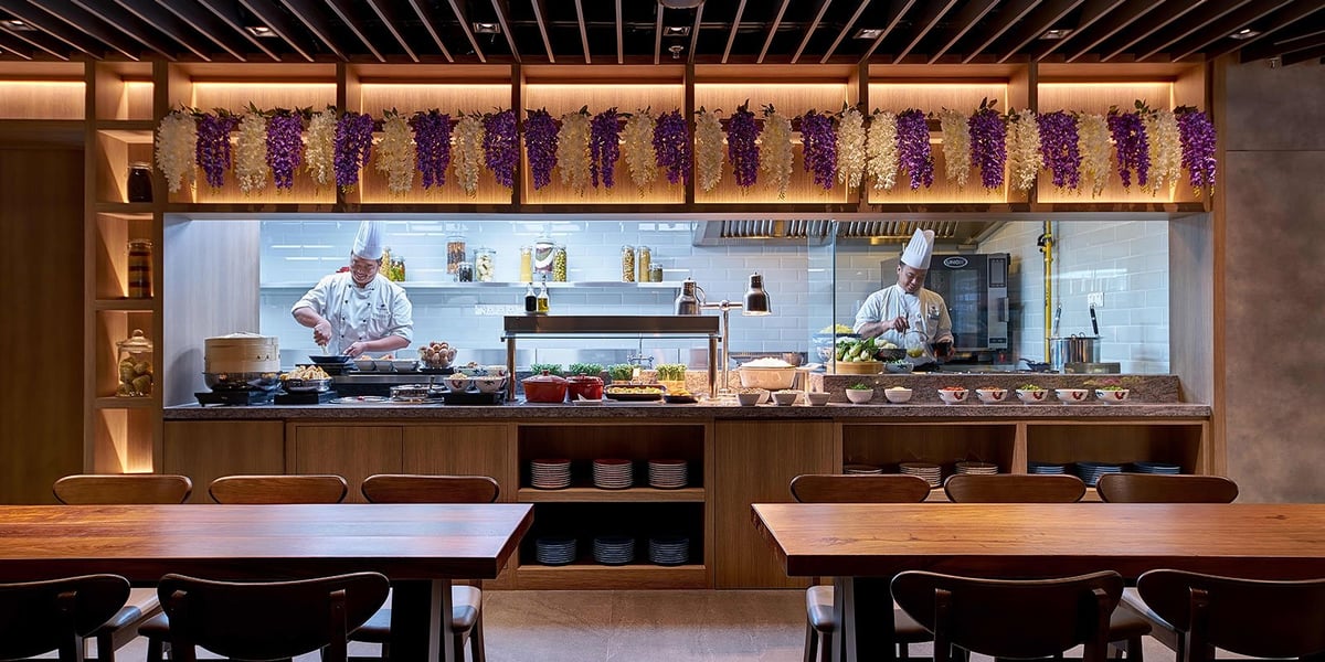 Chefs preparing fresh meals at the live cooking station in the Plaza Premium Lounge at KLIA Terminal 2, with hanging floral decor, wooden counters, and a selection of local dishes and ingredients on display.