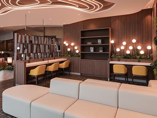 Stylish co-working and lounge area in the Travel Club Lounge at KLIA Terminal 2, featuring cream-colored seating, yellow-accented work chairs, decorative lighting, and modern ceiling fixtures with curved designs.