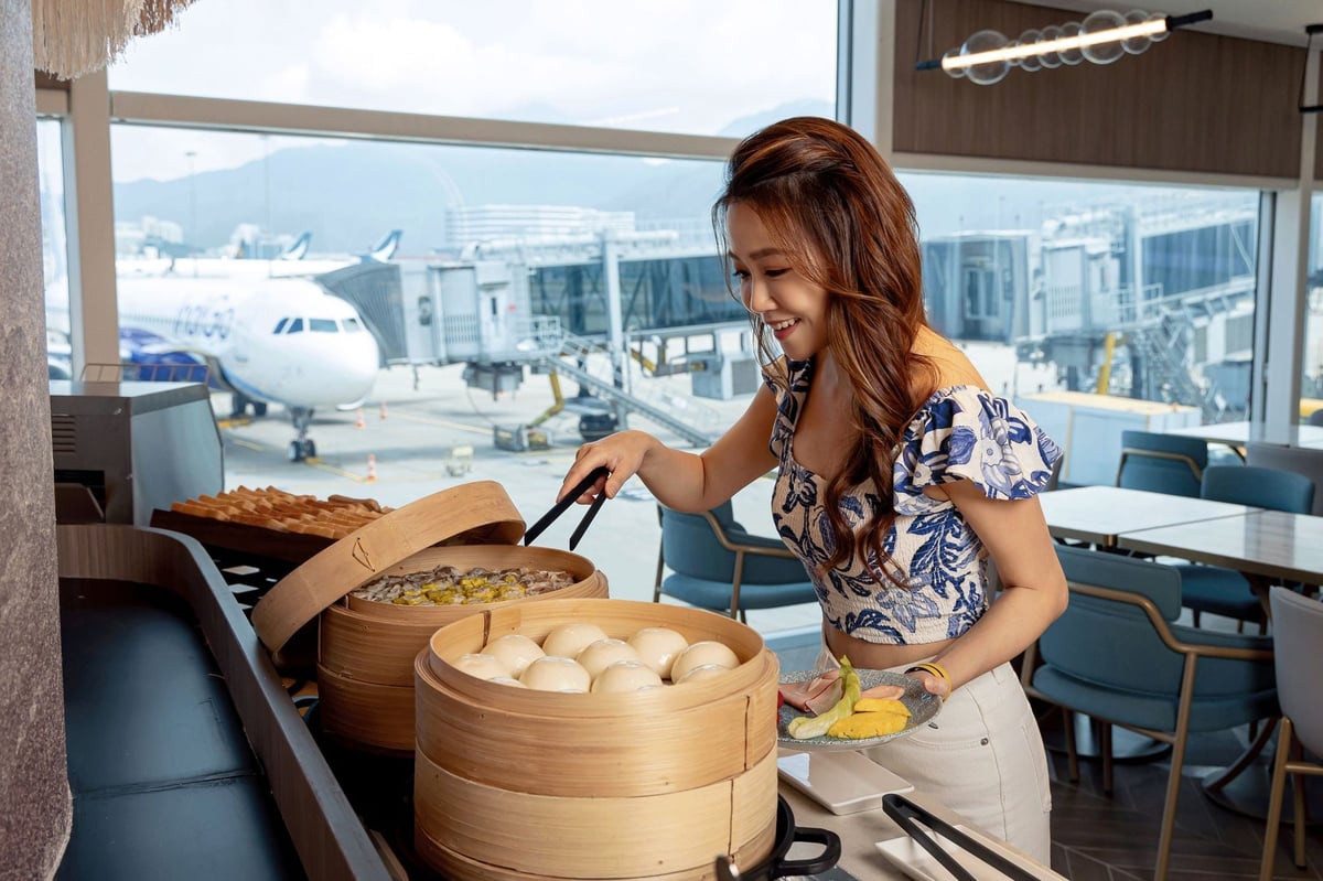 A smiling woman in a blue and white floral top serves herself dim sum from bamboo steamers at the Kyra Lounge buffet at Hong Kong International Airport, with large windows offering a clear view of the tarmac and parked aircraft in the background.
