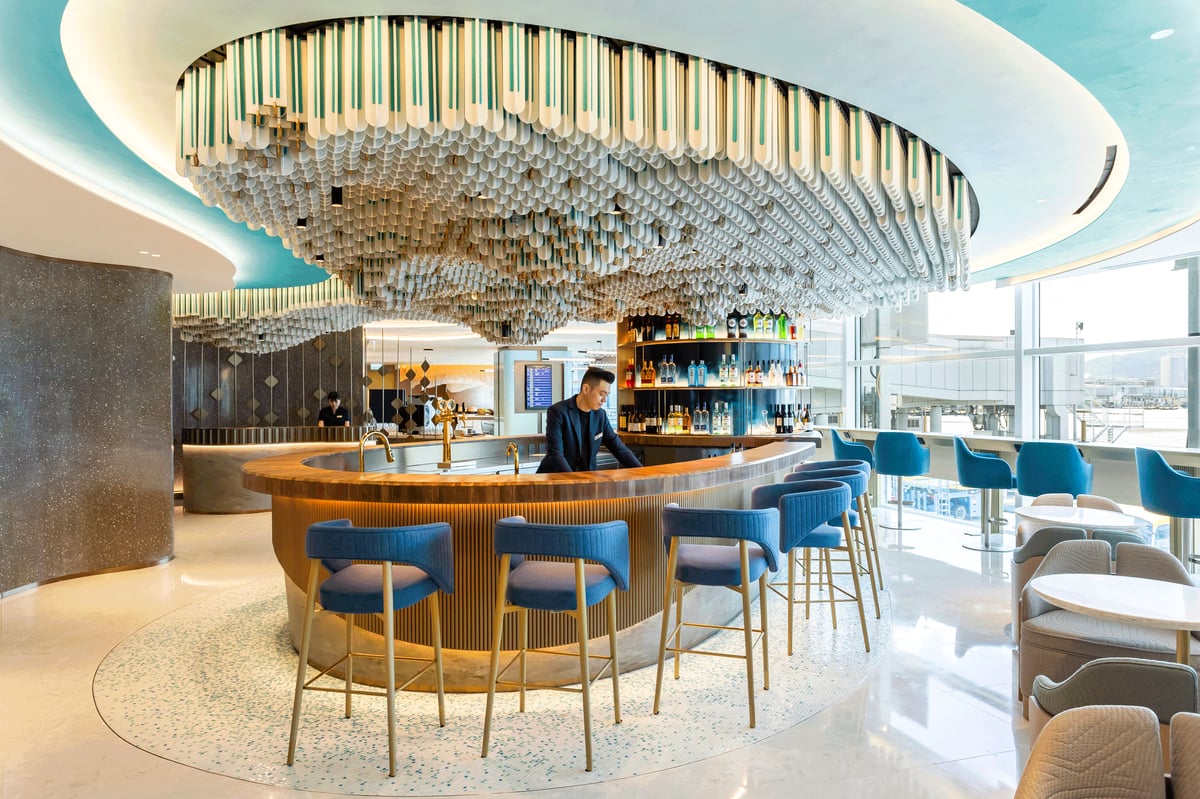Stylish bar area inside the Kyra Lounge at Hong Kong International Airport, featuring a circular wooden bar counter with blue upholstered bar stools, a backlit display of premium spirits, and a dramatic sculptural ceiling installation.