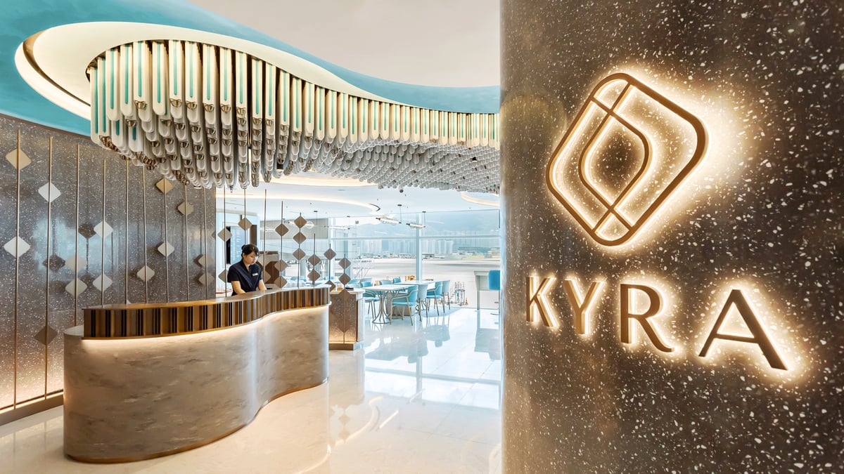 The entrance to the Kyra Lounge at Hong Kong International Airport showcasing a modern curved marble reception desk beneath a chandelier installation, with a glowing Kyra logo illuminated on a speckled stone wall and views of the bright, airy lounge.