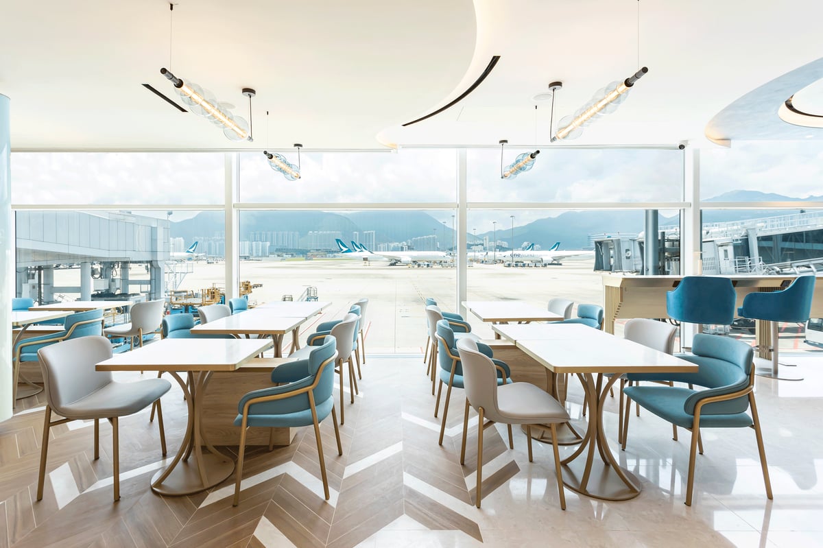 Bright and airy seating area inside the Kyra Lounge at Hong Kong International Airport, featuring modern white and blue chairs, light wood tables, and large floor-to-ceiling windows.