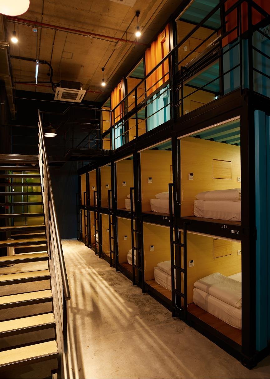 Industrial-style sleeping pods at CapsuleTransit in KLIA Terminal 2, arranged in a stacked layout with black metal frames, light wooden interiors, ladders for upper pods, and a minimalist concrete floor under warm, ambient lighting.