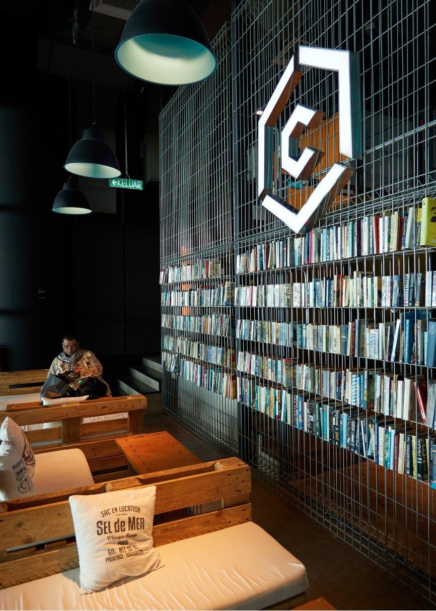 Cozy reading corner at CapsuleTransit KLIA Terminal 2 with wooden pallet seating, white cushions, and a tall wire-grid bookshelf filled with books, all set under soft industrial pendant lighting and a minimalist black interior.
