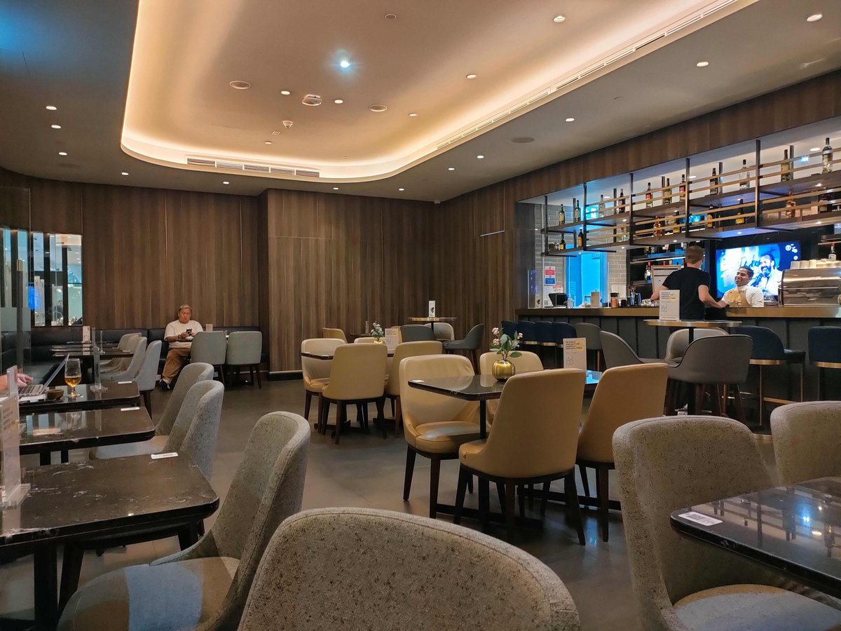 The image shows a stylish bar and dining area in the Plaza Premium Lounge in Dubai, featuring modern decor with sleek wooden panels and soft lighting. Comfortable chairs and tables are arranged throughout the space, with a few guests relaxing.