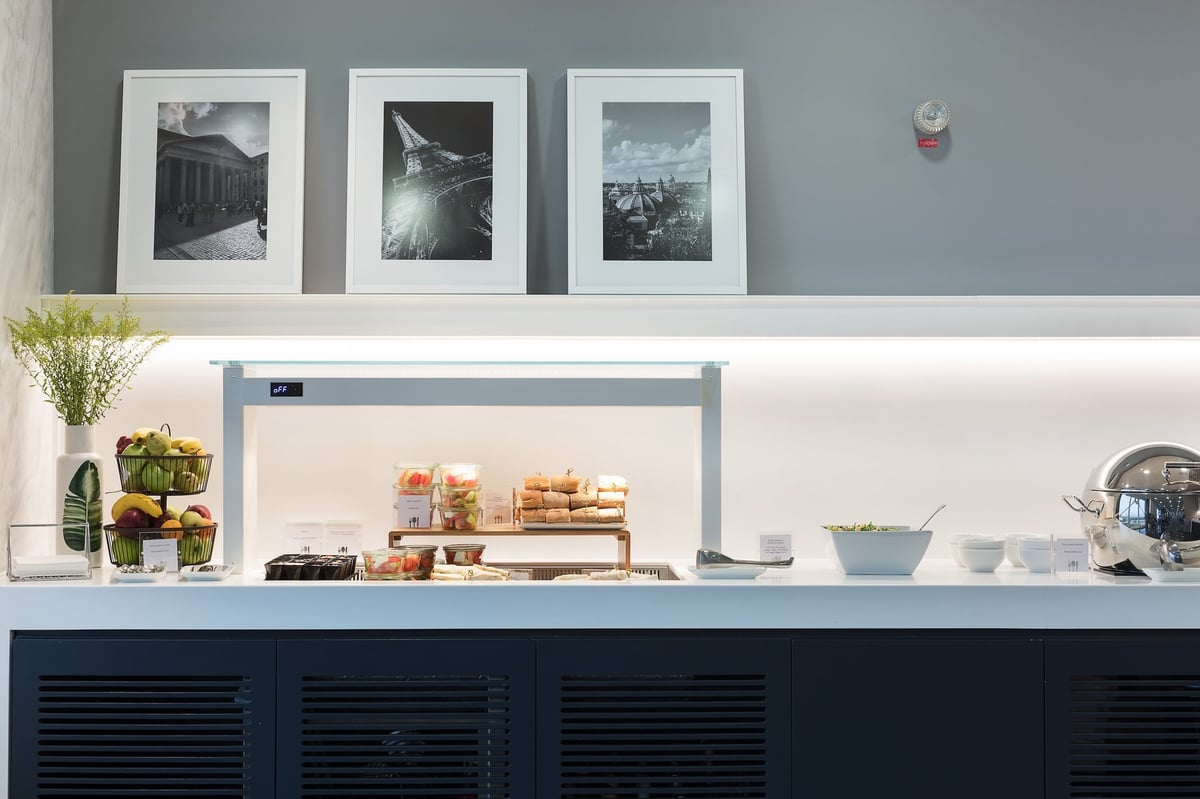 Buffet section at the Goldair CIP Lounge at Athens International Airport featuring a variety of fruits, breads, and salad food items on display. There are three black and white photographs on the top wall of the buffet and a decorative plant at left.