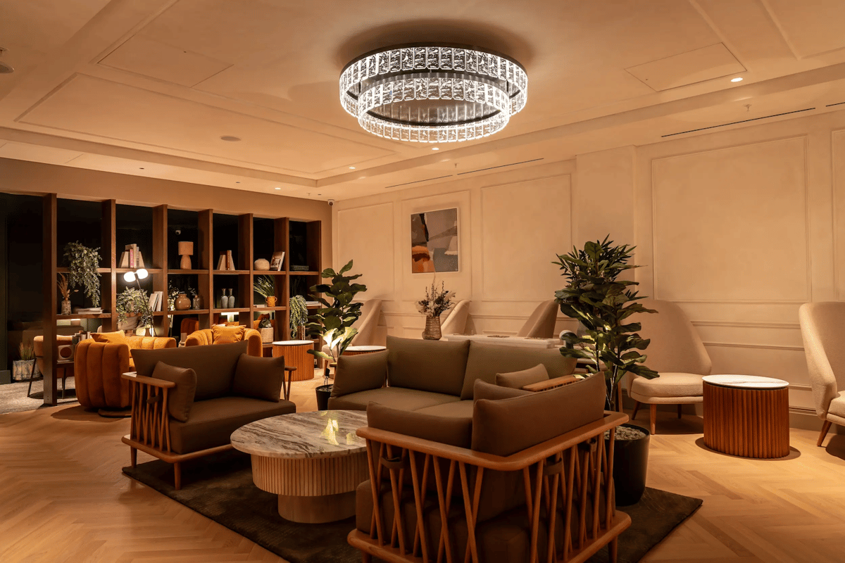 No1 Lounge at London Heathrow Airport featuring a dimly lit interior with shelving in the background and plush green sofas in the foreground. Two plants sit at the back of the chairs. Along the far wall are more armchairs in grey.