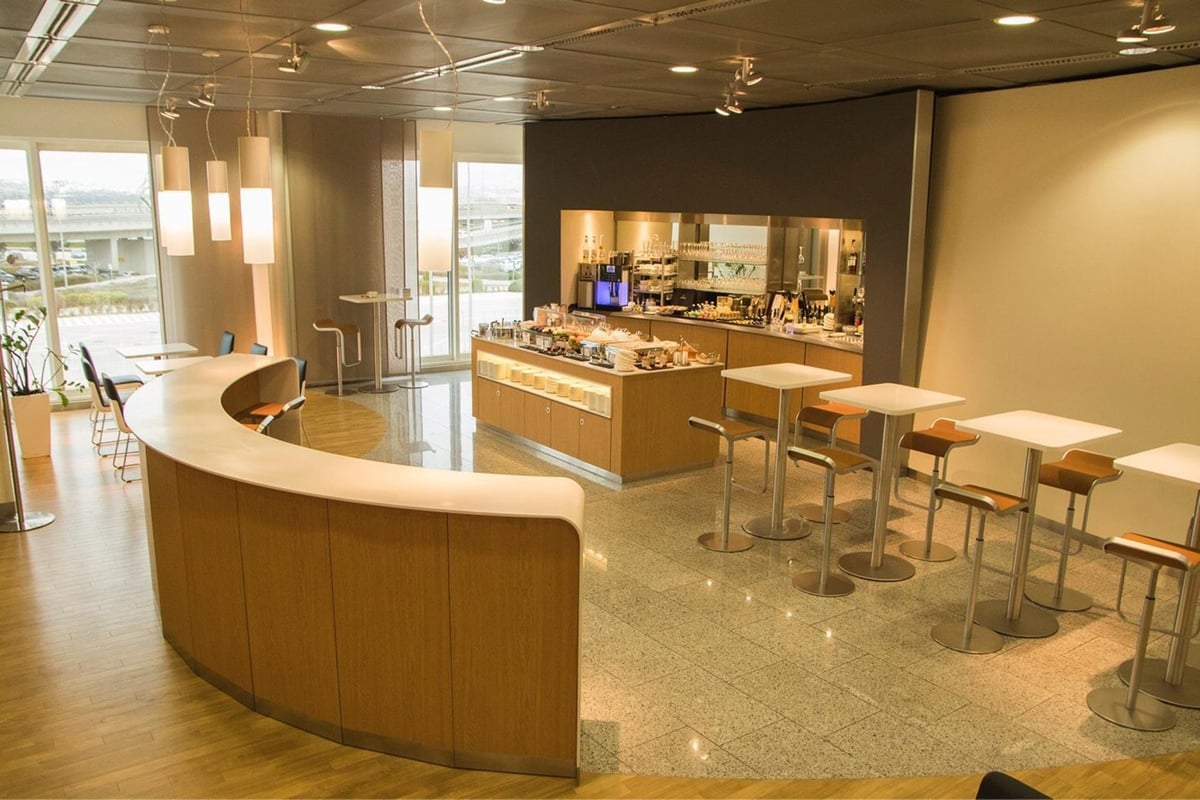 Warm and welcoming interior buffet and dining section of the Lufthansa Business Lounge at Athens Airport. The design is natural wood and curved finishes. Sleek bar stools line the tables, and warm lighting illuminate the space.